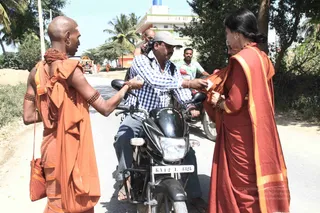 A street scene on a sunlit paved road with dirt verges, palm trees and a multi-storey building behind. In the foreground a seated man in a blue checked shirt and cap, on a black motorcycle with registration KA42L1825, accepts a folded paper from a woman in a rust-red sari with orange-yellow borders. To the left a shaven-headed monk in orange robes and rudraksha bead necklaces and bracelets reaches toward him, holding a small orange cloth bag. Behind them, further orange-robed figures walk near a decorated vehicle with orange flowers, and another man in a grey shirt rides a scooter.