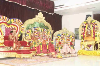 Indoor temple-style ceremony on a white tiled floor before a maroon velvet drape. A man seated at left on a red velvet and gold throne-like seat, wearing a red-brown cap, orange-red garments and beaded necklaces, holds his palms together in a greeting. Three deity murtis sit on a central golden peetha draped in garlands of red, yellow and white flowers, with fruit offerings and plates of food spread on the floor before them. To the right stands another garlanded murti on a golden seat, and a further golden throne with flower garlands is empty. Brass pots and a flower-decorated altar complete the setting.