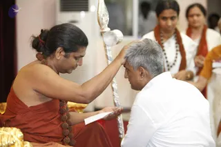 A man in a saffron-orange dhoti and rudraksha mala sits cross-legged and places his right palm on the head of a grey-haired man in a white shirt who kneels before him, a blessing gesture. The seated man has long hair tied in a knot with a red band and a rudraksha mala draped over his bare shoulder; he holds a folded white paper in his left hand. Behind them, two women in white with orange shawls stand, one wearing a long beaded necklace. A silver ceremonial staff with a finial stands beside the seated figure, and gold cushions appear at the lower left. The setting is an indoor hall with a white air-conditioning unit on the wall. Three people are visible. The seated figure is an appearance-based probable Swamiji candidate, identified by the long hair, rudraksha malas and saffron clothing, and is physically present.