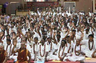 A large crowd of men seated on the floor in a hall, most dressed in white shirts and white dhotis with rudraksha malas, raising their right hands high in unison. In the foreground at left, a shaved-headed monk wears an orange-saffron robe with rudraksha beads and a bangle. Behind them a banner reads Nithyananda Galleria above shelves of items, and a wall displays framed images. Yellow papers rest on the mats in front, and a microphone stand is visible at center.
