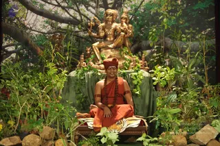 A seated person in saffron-orange robes and an orange turban sits with eyes closed on a wooden seat draped with a tiger-striped cloth, in a lush artificial garden of green plants and rocks. Behind, a large golden deity murti with multiple arms sits on a green-draped pedestal framed by a painted backdrop of a great tree, with smaller golden figures and green garlands around it. A rudraksha mala hangs over the chest and bare arms bear orange armbands. The seated person has long braided hair and is in a meditative posture with hands resting on the lap.