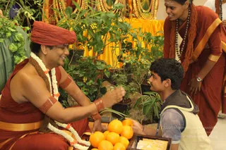 A seated man in saffron-red robes and a red turban wears a white flower garland and rudraksha mala, with a gold-banded waist cloth and red beaded armbands. He smiles and reaches toward a potted plant among green foliage, while a young man kneels in a grey hooded vest and blue lanyard, holding a tray of oranges. Behind them a standing man in red-orange robes and a rudraksha mala looks on, and a golden altar cloth with garlands is visible in the background.
