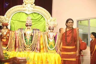 A photograph in a hall with a cream wall and glass windows. Three deity murti stand on a golden arched throne with a green backdrop, decorated with garlands and jewelled crowns. A man in an orange-red robe with a long rudraksha mala and a red bindi stands at the right, looking to his left. Behind him, other people in orange and saffron robes stand nearby, and a woman in white is at the far right. Yellow cloth covers the platform beneath the deities.