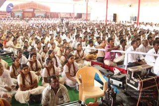 A large photograph of a satsang audience seated on green mats under a open-sided pavilion with red pillars. Hundreds of devotees fill the floor, many women in white saris with red-orange shawls and rudraksha-style necklaces, and men in white shirts and dhotis, some in pink or striped shirts. In the foreground a single empty beige plastic chair faces the crowd beside a red-draped platform and audio equipment with cables and a camera case. Video cameras on tripods stand at the back, and a distant stage structure with a banner is visible. No a person candidate is visible in the frame.