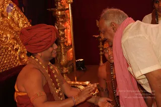 A photograph in a red-curtained shrine room beside a golden throne. A figure in a red-orange turban, gold-beaded necklaces and a red-orange wrap is seated on the left, bending forward and extending both hands to receive or offer something from a golden tray. Facing them, an older man with white hair and round glasses wears a pink shawl over white clothing and leans forward with hands together. Behind them a bare-chested attendant with a sandalwood-marked forehead and rudraksha mala stands by a brass lamp stand, and another attendant in white stands at the upper right. Gold ornamental carving is visible on the throne. Two or three people are visible.