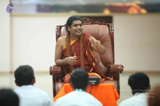A man with long swept-back dark hair sits on an ornate carved wooden throne with brown leather upholstery, gesturing with his right hand while holding a gold pendant. He wears a rust-red and saffron shawl over bare shoulders, with a long string of brown rudraksha beads around his neck and a bracelet on his wrist. Several people in white clothing sit with their backs to the camera in the foreground, facing him, with a microphone and a Yamaha device on an orange-draped table. The setting is a bright indoor hall with cream walls. a person figure with distinctive long hair, saffron-red garments and rudraksha malas is seated as the speaker, giving a talk to the audience.
