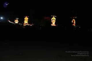 A night photograph of illuminated processional vehicles in a dark open ground. Four ornate lit floats or chariots with canopies, glowing with lamps and decorations, stand in a row; the nearest carries figures under a golden canopy. Strings of multicoloured lights trail along the left side. No people are clearly visible, and no one is identifiable as a person. The lower half is almost entirely dark ground.