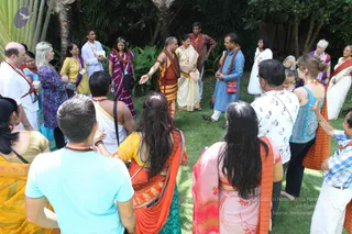 Outdoor photo of a circle of devotees on green lawn beneath tropical trees and palms. A bare-chested man in orange-and-maroon dhoti-style wrap with rudraksha-style beads stands centre-left with arms outstretched, beside a woman in cream and gold sari. Around them stand a man in a light blue kurta, a woman in purple and blue patterned sari, a man in white with a sash, and many others in saris, salwar and casual clothes, some wearing lanyards and orange-red shawls. Some onlookers have their backs to the camera, including a woman in orange sari and a woman in pink sari. The bare-chested central figure shows distinctive adornments but his identity is uncertain from appearance alone.