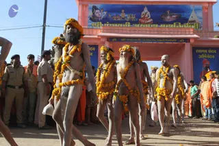 Outdoor procession of naked sadhus (Nirvani akhada ascetics) walking forward through a crowd, bodies smeared with ash, wearing marigold garlands across their chests, shoulders and waists. Several have long white or grey beards and long hair. Uniformed police officers in khaki stand at left and watch the crowd. Behind them an ornate pink gateway bears a painted banner with Shiva, a seated saint and temple imagery, with the lettering Shri Panchayati Akhara Mahanirvani. Another banner reads Swagat Vandan Abhinandan. Devotees in orange and saffron clothing and onlookers in casual clothes line the path under a clear blue sky.