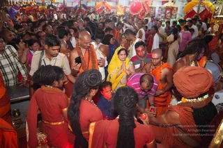 A dense crowd fills a temple or camp interior at a festival, with many people in saffron and orange robes and red sannyasi-style garments. In the foreground, a bearded sadhu in orange wrap and rudraksha beads sits at the right, while a woman in a yellow sari with folded hands stands centre, and a man in a pink shirt bends forward. Several bald-headed men wear orange cloths and garlands, and red umbrellas and yellow decorations hang overhead. Devotees press close, some holding phones and red flowers. Several priests in orange robes and rudraksha malas stand among the devotees, and a man at centre left wears a yellow-green check shirt beside a uniformed official with a lanyard. No single figure can be confidently identified as a person from appearance alone.