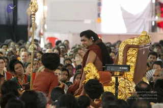 A photograph of a large seated crowd in a hall. A man with long dark hair, wearing a rust-red shawl over bare shoulders and a headset microphone, sits on an ornate gold-and-brown throne with carved gold armrests and a Yamaha keyboard unit beside it. Near the throne stands a man in orange-red clothing and a dark head wrap, facing him, and a gold staff with an ornate top is visible at left. Many devotees in saris and kurtas, some with orange and white garments, fill the foreground and background.