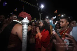 Night photograph of a crowd at an outdoor religious gathering lit by a bright overhead lamp and handheld phone lights. In the centre, a bearded man with long grey-streaked hair wears an orange-red robe and presses his palms together in namaste, facing a man seen from behind with long dark hair, a red turban and a white flower garland, who holds a tall silver staff with a finial. To the right, a young man with a shaved head wears an orange robe and gold-beaded necklaces, with a red tilak on his forehead. Other devotees in red and maroon robes and white forehead markings stand nearby, and saffron flags fly in the background. Several people hold up phones to record.