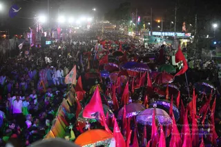 A dense night-time street procession crowded with many people, lit by bright white floodlights and strings of lights along the road. Numerous pink, red and orange flags flutter above the crowd, and many decorated umbrellas in purple, pink and orange with sequined trim are raised over the marchers. Some people stand on raised platforms or ladders at left, while colourful banners and signboards line the route. The scene conveys a large festival procession moving through a city street, with tall ornamental structures visible in the background.