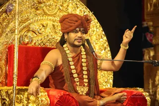 A man seated on an ornate gold throne with red velvet cushions, wearing a rust-orange turban with a vertical tilak on his forehead, an orange-saffron shawl over the shoulders, a heavy gold bead necklace with a gold pendant, multiple gold armlets and bangles, and a rudraksha-style brown beaded mala draped at the chest. He is speaking into a microphone on a stand while raising his right index finger in a teaching gesture, with the left arm resting on the throne. Probable Swamiji seated in the teaching position, with long swept-back hair and ceremonial gold jewellery visible. Gilded carved panels fill the background.