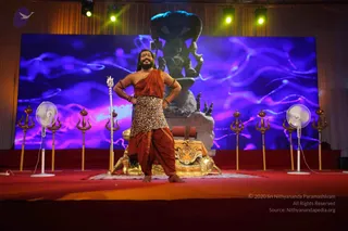 A photograph of a bearded man standing on a red stage in front of a large purple-blue video backdrop. He wears an orange-red shawl over his shoulders, a leopard-print tunic, and rust-orange dhoti-style trousers, with bangles on his wrists and a red bindi on his forehead. He stands with his hands on his hips and smiles. Behind him is a grey stone statue of a deity with a multi-hooded cobra canopy, flanked by a golden tiger throne draped with an orange cloth. A tall trident stands at his side, and several golden tridents line the stage, with two white pedestal fans in view. a person: a bearded man in orange and leopard-print clothing with bangles and a bindi, standing as the central figure on stage.