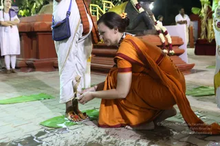A woman in an orange sari with a maroon border kneels on a paved courtyard at night, her hands touching the bare feet of a man standing in white dhoti and shawl with an orange border. Both feet rest on green leaves, and red kumkum marks are visible near the toes. Her hair is pinned up with an orange flower. Behind them, a stone structure is decorated with a garland of white flowers and red beads, and a potted plant stands at the right edge. Other people in white clothing stand in the background.