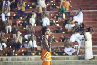 Outdoor evening scene on a red stone stepped tank at dusk, with many seated people in white and bare-chested devotees with oil lamps set along the steps. In the foreground a young boy stands with palms pressed together in namaste, wearing an orange dhoti with yellow border, a rudraksha mala, and white markings on his chest and forehead, gazing upward. Behind him rows of seated figures in white and colourful clothes hold lamps, and a cameraman with a video camera on a tripod films on the right. Small brass oil lamps glow across the steps.