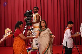 A photograph of a stage set with a deep red draped curtain and a red floor. A cameraman in a white dhoti with gold border and a headset stands behind a tripod-mounted video camera. In the foreground, a woman in an orange sari holds a wooden tray with a framed picture of a seated saffron-clad figure. In the centre, a woman in a cream sari with green and orange borders, a necklace and red bindi gestures with an open hand. On the right, a person in a white kurta with an orange shawl holds a brass tray with a lamp. A decorative colourful cloth lies on the floor by the camera.