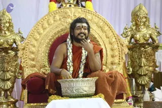 A seated man in saffron-orange robes sits on an ornate gold throne with a red cushion, flanked by two large gold female figures holding lamps. He wears a long rudraksha-style mala of white and blue beads with white flower garlands, a red bindi and white forehead markings, and a neatly trimmed beard. His right hand rests near his chin as he smiles, and his left hand rests on a large silver bowl filled with yellow flower petals. A white-draped table and a dark red cushion sit below. A small purple logo appears at the upper left, and a faint copyright line is visible at lower right.