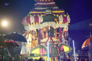 A night photograph of a festival procession with a tall, elaborately decorated palanquin or ratha covered in garlands of yellow, orange, purple and white flowers, with a green leafy canopy and a tiered top. A man in a saffron-orange garment with a gold crown and rudraksha-style beads sits seated on a gold-trimmed throne at the centre of the vehicle. Around him, several men in white dhotis and shawls, one with a bare upper body and bead necklaces, stand on the platform and hold dark umbrellas and fans. Coloured umbrellas and bright white light poles frame the scene, with a crowd visible at the bottom edge.