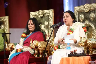 A photo of a stage event with two women seated on ornate gold-and-carved thrones with elephant-shaped armrests and carved face medallions. The woman on the left wears a magenta and blue sari with a red bindi and a white jasmine and red-and-yellow flower garland. The woman on the right, in a white embroidered top and white shawl with a similar garland, speaks into a microphone while holding a pen and a sheet of paper. A water bottle and a small table with an orange cloth sit in front. Deep red curtains form the backdrop. No a person candidate is visible; the thrones are empty of any figure in the frame.