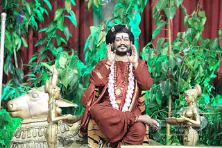 A seated figure in rust-red and maroon robes with gold-bordered shawl sits on a tiger-striped seat, hands raised palms outward beside the face in a blessing gesture. He wears a long garland of white and pink flowers, a rudraksha mala with a gold pendant, and a tilak on the forehead, with long dark hair swept back under a crown-like ornament. Beside him stands a golden bronze statue of a bull, Nandi, and a golden female figure holding a lamp. Green leafy bodhi-style foliage and a deep red curtain form the backdrop. Patterned green lighting washes the scene. A