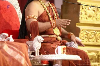 A seated figure on a red throne-style chair wears a rust-orange draped garment, layered gold-toned beaded necklaces, an ornate gold pendant, orange and white beaded armbands and bracelets, and a jewelled belt. Long dark hair falls behind the shoulders. A silver lion figurine and a silver ritual tray with a white mug sit on a silver table in front, with a microphone stand and gilded carved panels behind. The figure is cropped at the chest and face, so no face is visible. a person: the rust-orange saffron-toned clothing, long swept-back hair and multiple beaded ornaments with a gold pendant are consistent with the page's claim, and the figure is seated on a ceremonial throne in a discourse setting. A mug printed with a portrait is visible on the table, which is a depiction, not the person present.