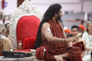 A seated man in rust-red saffron robes sits on an ornate silver throne with a bright red velvet cushion, holding a small golden trident-shaped ritual implement in his right hand. His long dark matted hair falls over his shoulders, and a white armband-style ornament is on his upper arm, with a red wristband. A copper vessel sits on the left of the throne. Behind him, people in orange and saffron clothing and white garments are seated at a event, and a yamaha keyboard is visible at lower left. Swamiji (probable): long dark hair, saffron robes and a ritual trident mark him as the seated central figure on the throne at a Navaratri darbar.