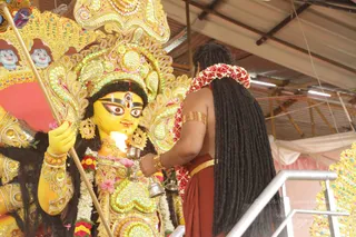 A photograph of a large, ornately decorated yellow-faced deity murti of Devi Durga, with a golden crown, gold jewellery and a gold-tipped staff held in its raised hand. In front of it, a man seen from behind, with long dreadlocked black hair and a red-maroon dhoti or wrap, wears a red and white flower garland and a gold armlet. He holds a silver lamp or bell-shaped puja vessel toward the murti. The setting is a covered hall with a metal roof, white cloth and pink draping, and a silver railing in the foreground.