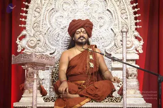 A man seated cross-legged on an ornate silver throne with lion-headed armrests and a leopard-print cushion, in front of red drapes. He wears a rust-orange turban with a small vertical tilak on his forehead, an orange-saffron draped garment, and a rudraksha mala with gold pendants and a gold necklace. A microphone stands beside him as he sits with eyes closed and hands resting on his knees. Probable Swamiji by appearance cues: orange turban, saffron clothing, rudraksha mala and gold ornaments, seated on a throne.