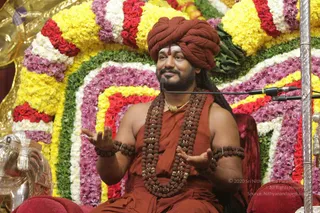 A seated man in a rust-orange turban and orange robe speaks into a microphone on a stand, gesturing with both hands. He wears multiple strands of rudraksha beads, rudraksha armlets, and a gold pendant necklace, with a red tilak on his forehead. Behind him is a large floral arch of yellow, white, red and purple flowers. A red cushioned seat and a silver ornamental animal figure are visible at the lower left. One person is visible.