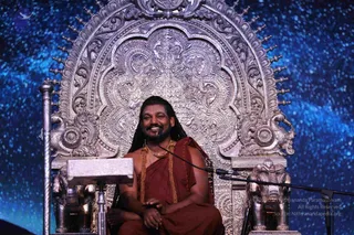 A man with long dark hair and a beard sits smiling on an ornate silver throne with intricate repoussé carvings, peacock motifs and finial spikes, flanked by silver elephant-shaped armrests. He wears a reddish-maroon robe draped over one shoulder, a gold necklace and gold beaded mala, and holds his hands together in his lap. A microphone stand stands before him beside a stone-top lectern, and the backdrop is a starry blue night sky. a person: the seated man wears long hair, a gold necklace and mala cues with a maroon robe, presented on a throne.
