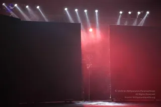 A dim stage lit by overhead spotlights and a red haze, with two large dark black panels flanking a central opening. In the middle stands a tall metal trident, a trishul, on a stand, partly obscured by smoke. The floor is dark and reflective. No people are visible in the scene.