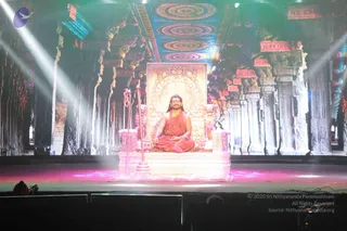 A seated man in orange-saffron robes sits cross-legged on an ornate golden throne on a stage. He wears a rudraksha mala with a long necklace and has long hair swept back. A tall staff with a trident-like top stands beside the throne. Behind him a large projected backdrop shows carved temple pillars and a colourful ceiling with circular patterns, lit by white spotlights. Several people are seated in the dark audience area at the bottom edge, and a camera operator is visible at the front left.