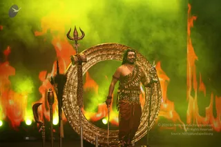 A performer on a stage stands before a large ornate golden ring-shaped prop, its surface carved with patterns, while orange and green flame-like backdrops glow behind him. He wears a bare chest with a leopard-print garment, a long reddish-brown lower cloth, layered metallic ornaments on the shoulder, and wrist bands; his hair is long and swept back, and he holds a trident-topped staff in his right hand. Beside the ring stand a tall trident, a curved ceremonial staff, and a sceptre-like rod with a tassel. Fog and coloured stage lighting fill the scene.