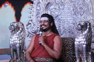 A photograph of a smiling man seated on an ornate silver throne with a leopard-print cushion, flanked by two silver lion statues. He holds a handheld microphone to his mouth while speaking, with his head turned slightly to the side. He wears a rust-red sleeveless garment, a leopard-print waist cloth, a gold pendant necklace and a red wrist thread, and has a white horizontal band on his forehead with long dark hair. The background shows a dark stage with flame-like decorative panels. a person (long swept-back hair, ornate gold necklace, distinctive clothing) seated in discourse.