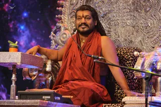 A seated man in a rust-red saffron shawl sits on an ornate silver throne decorated with carved floral and figural relief, with a leopard-print cushion beside him. He has long dark hair swept back, a red tilak on his forehead, a gold pendant, and a red thread bracelet on his wrist. A microphone stands near him, and his hand rests on a silver lectern with elephant-head supports. A water bottle sits on the lectern and a cosmic blue-purple backdrop fills the background. Swamiji (probable): the long swept-back hair, saffron shawl, gold pendant and ornate silver throne mark him as the presenting teacher, giving a discourse on stage.