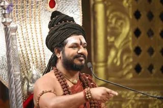 A man with long dreadlocked hair wound into a high topknot decorated with rudraksha beads sits at a microphone on a stand, speaking and gesturing with his right hand pointing to the side. He wears a red-orange saffron wrap over bare shoulders, with large rudraksha mala necklaces, a gold pendant necklace, rudraksha armlets and a bead bracelet. A vertical red-and-white tilak marks his forehead. Behind him are ornate gold carved panels and a silver throne with decorative chains. a person is shown in the setting of a public satsang discourse; the visible cues are the dreadlocks with rudraksha, saffron wrap, rudraksha malas and gold ornaments, and he is seated before a microphone as the speaker.