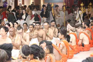 A large seated congregation fills a ceremonial hall during a ritual. Foreground devotees in orange and saffron cloths sit on the floor, some with hands pressed in anjali, others with eyes closed. Several men in white dhotis and bare upper bodies, with rudraksha malas, sit with palms joined, one with a red-and-white tilak. Women wear orange garments and hair buns with flower decorations. Behind them, rows of people in white and red clothing sit on white chairs. Two video cameras on tripods stand among them, and gold ornamental finials decorate the room's edge.