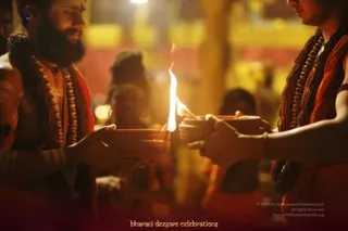 Two men in saffron-orange robes hold clay oil lamps with lit flames at a night ritual, with the flames rising between them. The man on the left has a full dark beard, smiles, and wears a dark rudraksha mala and a long gold-coloured bead necklace over his bare upper body. The man on the right, shown partly cropped, wears a similar brown bead mala and orange cloth and holds his lamp with both hands. Blurred figures and warm golden lights fill the background.