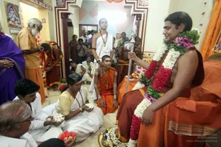 A seated figure in orange robes with long swept-back dark hair, wearing layered garlands of white jasmine, red and pink flowers and green leaves, holds a silver staff with an ornate top and sits on an orange-draped seat in a decorated room. Before him, a young boy in orange robes and rudraksha beads kneels, and a young man in a white dhoti stands with a sacred thread. A woman in a yellow-bordered white sari sits on the floor offering white flowers, a man in white holds flowers, and a seated elderly man in orange with a white beard watches. A brass plate of fruit and flowers rests at the front. Pink and red flowers and small figurines are visible on a side table, with a painted deity image on the wall.