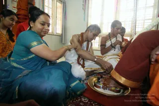 A photograph of a domestic room during a padapuja ritual at a house visit. A woman in a teal sari with gold border sits on the left, resting her hand on a man's arm. A man with grey hair, bare-chested and wearing a white dhoti with gold border and a rust-coloured rudraksha mala, kneels and washes the feet of a person in a maroon garment with gold border, with a brass plate holding offerings beside them. Another person in a white dhoti and rudraksha mala sits behind, and a young woman in orange stands at the left edge. Sheer blinds and a pale wall frame the room.