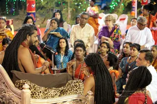 Evening outdoor satsang on a lawn strung with green fairy lights and a red-and-blue banner. A bearded man with long dreadlocks, bare-chested with an orange-red shawl, sits on an ornate carved pink-wood throne draped with a leopard-print cushion, speaking into a microphone on a stand and gesturing with his right hand. Beside him, a young devotee with long dark hair and an orange shawl and beaded mala holds a small object and listens. In front, a woman with long dreadlocks in orange sits facing him. Behind them, a seated audience of men and women in saris, kurtas and shirts fills white plastic chairs, several holding small plates, with a woman in orange at the right edge.