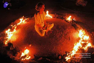 A seated figure in white clothes sits cross-legged at night inside a circle of burning fire, with a rudraksha mala hanging around the neck and hands resting in the lap. The ground inside the ring is smooth, with circular raking marks in the sand. Flames of varying height ring the seated figure on all sides, glowing orange against the dark surroundings. The person has shoulder-length light hair and faces slightly away from the camera, eyes closed in meditation.