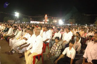 Night outdoor gathering of a very large crowd seated on red plastic chairs and on sand in rows facing a stage. Most attendees wear white shirts and white dhotis; some wear checked or plaid shirts and a few older men wear pink or red headcloths. Strings of bunting flags stretch across the open ground, with floodlights and a tall illuminated red figure of a deity murti in the background, plus a large portrait banner on the left. Palm trees and trees frame the scene.