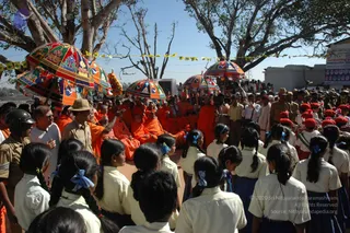 A festival procession outdoors beneath large shade trees with a bright blue sky. Several saffron-robed sannyasis and monks sit or stand in a group at centre, many with shaved heads, one in a saffron shawl gesturing with an outstretched arm. Ornate multicoloured parasols with embroidered panels and tassels are held aloft above them. In the foreground, rows of schoolgirls in white blouses and navy skirts wear blue hair ribbons, with a crowd of onlookers behind them, including khaki-uniformed police or officials and men in white. Bunting strings hang between the trees, and a building with signboards stands at right.