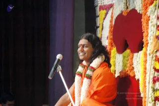 A smiling person in an orange-saffron robe sits beside a microphone on a stand, facing left, on a stage decorated with garlands of white, yellow, orange and red flowers and a large red felt cutout shape. The person has long dark hair, a red bindi and a white and green-striped flower garland around the neck. A dark purple curtain hangs at the left edge of the stage. The scene suggests a satsang or discourse. Swamiji (probable): long swept-back hair and saffron robe with flower garland, in a speaking role at the microphone.