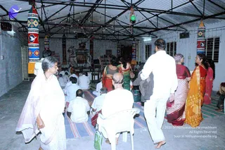 Interior of a covered hall with a corrugated metal roof and string lights, where devotees gather for an evening event. In the foreground, a woman in a white sari with a light shawl stands at left, and a bald man in white shirt and trousers sits on a white plastic chair. Several people in white kurta-style clothing sit cross-legged on striped mats facing a raised altar at the back, which holds a deity murti and a decorated throne-like seat. Women in saris of red, green and orange stand at right. Tall decorative fabric banners with geometric patterns and dove motifs hang along the walls. Several people, roughly 21-100 in total, are visible.