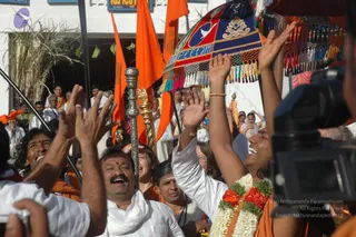 A joyful procession scene in front of a white building with orange flags and tall staffs. In the foreground, a smiling man in white clothes and a white shawl raises his hands, beside a man with a moustache and red tilak, and a woman in white with a white and red-green flower garland raising her arms. Behind them, devotees in white and orange clothing gesture upward, and a decorated canopy with red, blue and gold fringes and a bird emblem is held overhead. A video camera is visible at right. Several dozen people are visible in the background.