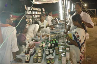 A night-time stall under a tent at an outdoor event, with bright white fluorescent tubes overhead. Shelves hold rows of books, CDs and DVDs, and a long table is crowded with bottled herbal remedies, jars and boxed products, some labelled Vilvanna. Several people in white clothes work the stall, including a woman in a white sari with a red-trimmed border and a mala, a man in white shirt and glasses, and a woman in a floral-patterned sari with gold jewellery who browses a yellow leaflet. A young man in an orange-trimmed shawl stands at the left edge. No deity, throne or a person depiction is visible.